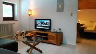 Eine Aufnahme des Wohnbereichs von Appartement 4 - "Schattberg" mit Sofa, Flat-TV und Blick auf den Esstisch in der Küche | © Haus Tannenblick